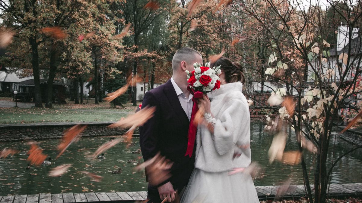 Newlywed couple kissing among falling leaves in a scenic autumn park.